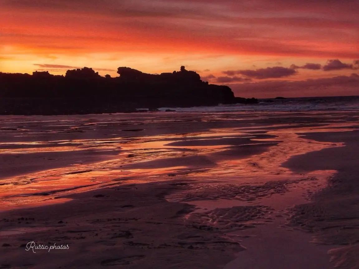 A serene beach at sunset with vibrant orange and red skies reflected in wet sand, silhouetted cliffs in the distance, and…