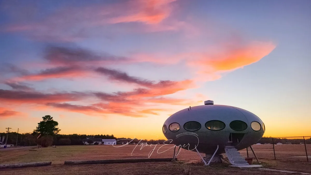 A silver, saucer-shaped UFO-style house with oval windows sits on stilts in a field during a vibrant sunset with pink…