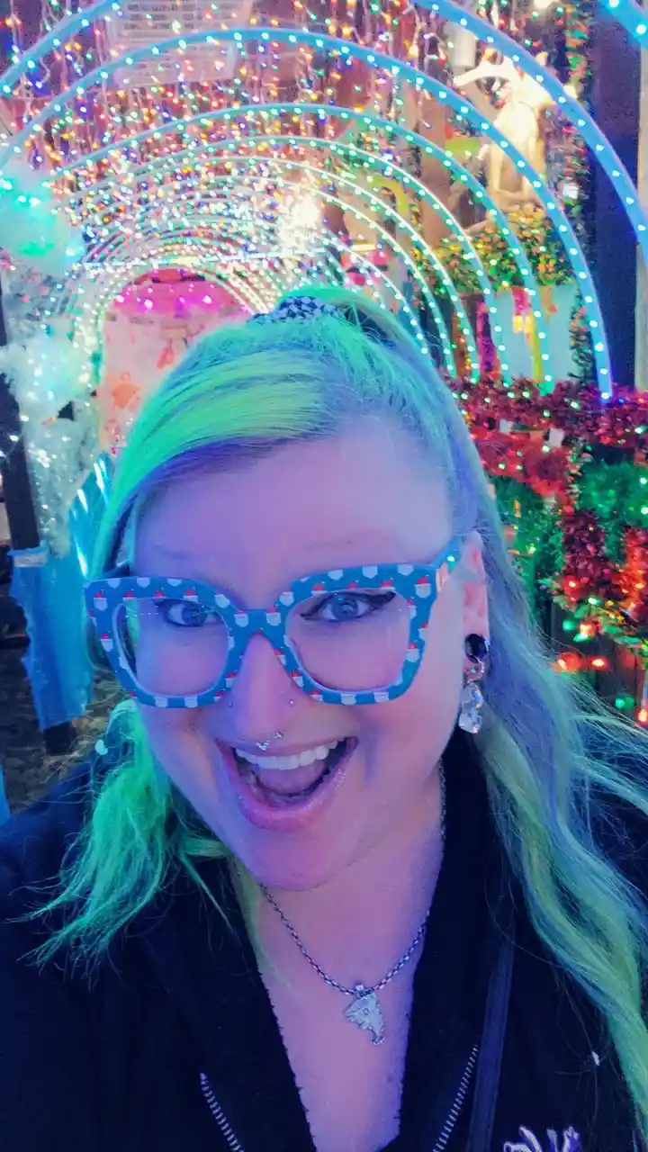 A person with bright green hair and festive blue glasses featuring Santa hats smiles widely at the camera. They are…
