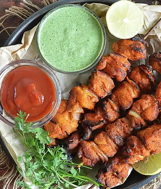 A top-down photo of grilled chicken tikka skewers arranged on parchment paper in a dark tray, accompanied by a bowl of green…