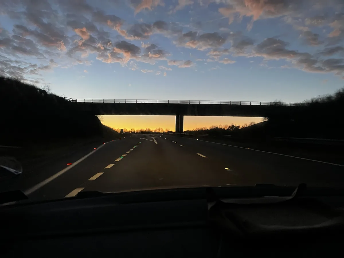A photo taken from inside a car looking forward on a highway at dusk, with a bridge overhead and a glowing orange sunset…