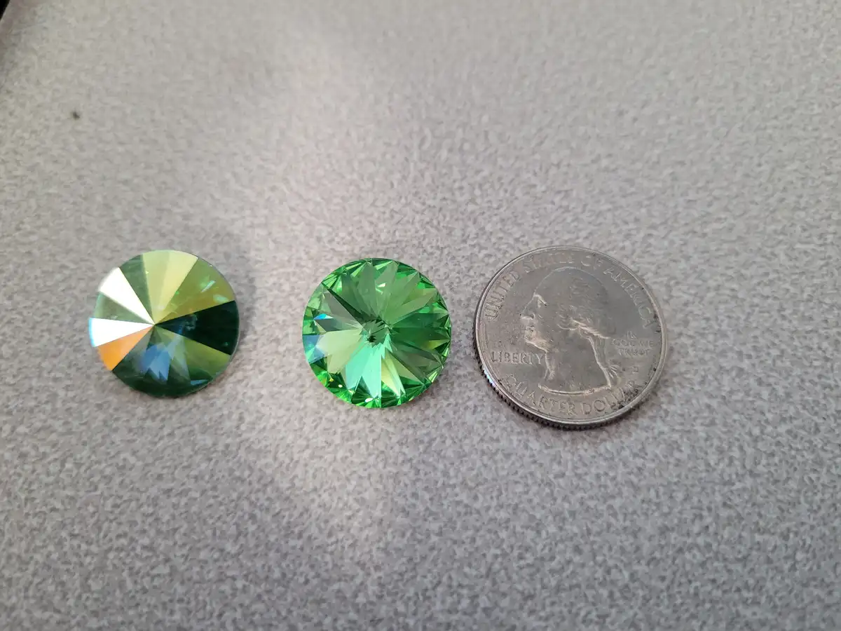 Three round, faceted green gemstones are laid out next to a U.S. quarter dollar coin on a speckled gray surface. The left…