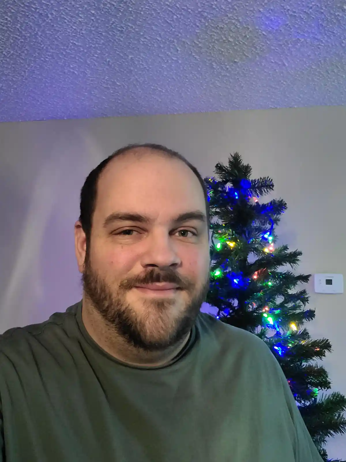 A man with a receding hairline and beard smiles at the camera, standing in front of a lit-up Christmas tree. The ceiling…