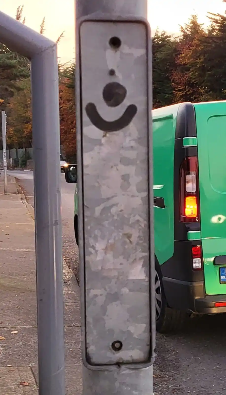 A close-up of a weathered, grey metal pole with a crudely drawn smiley face (two dots for eyes and a curved line for a…