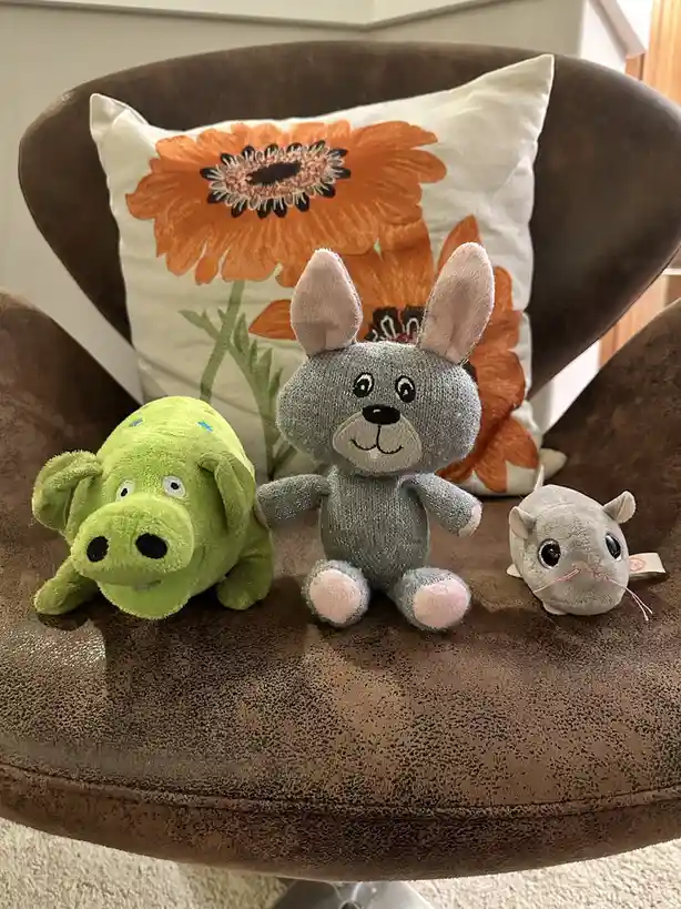 Three plush toys—a green pig, a grey rabbit, and a grey mouse—are arranged on a brown upholstered chair with a white pillow…