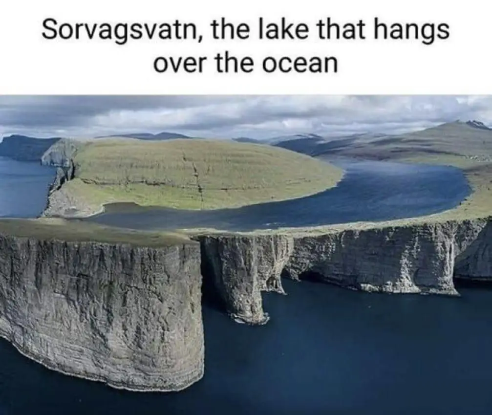 An aerial view of a dramatic landscape where a deep blue lake, identified as Sorvagsvatn, sits atop a steep cliff edge…