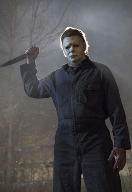 A still image of Michael Myers from the Halloween movie franchise, wearing his iconic white mask and dark coveralls, holding…