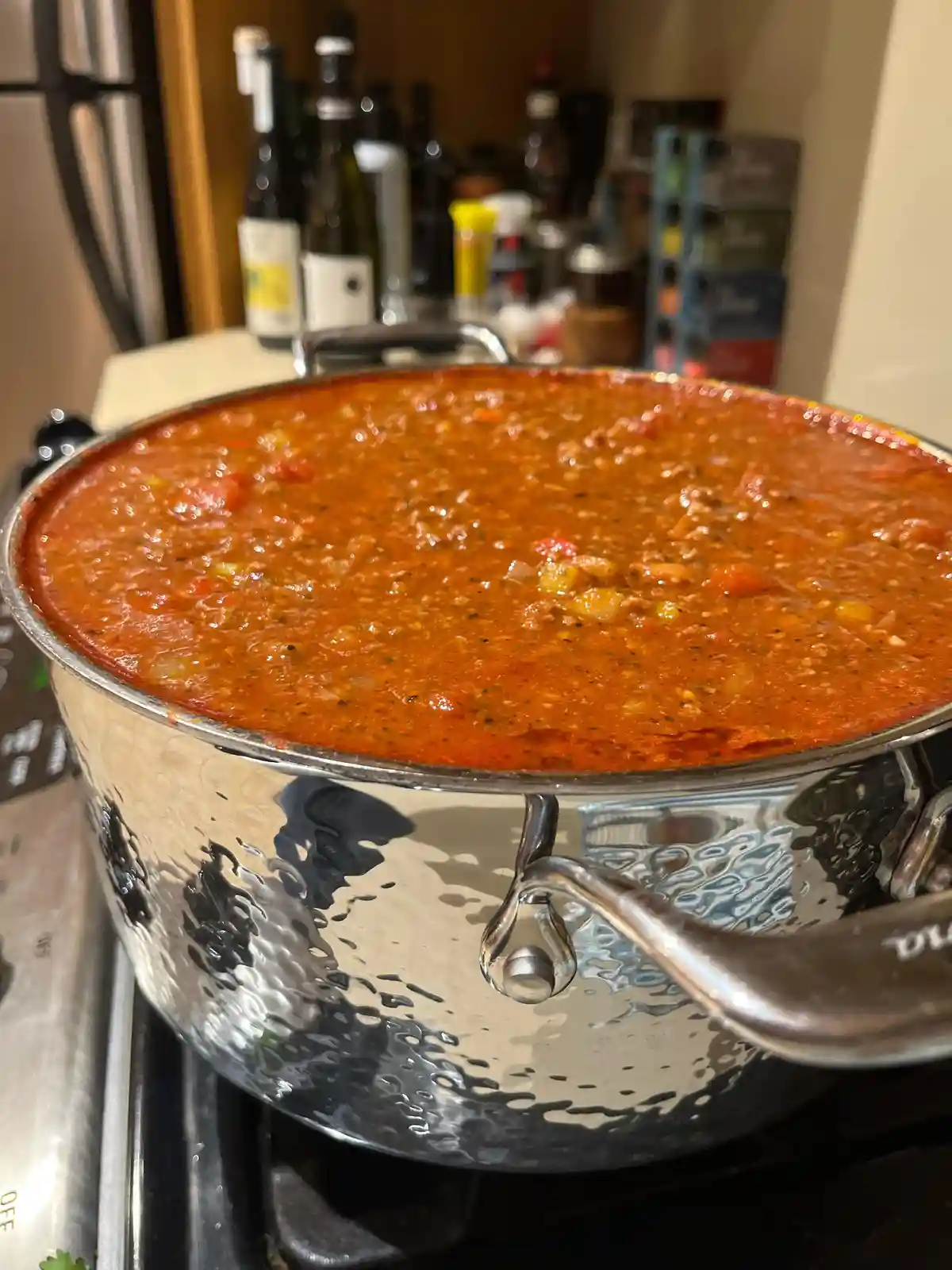 A close-up photo shows a large, hammered stainless steel pot filled to the brim with a rich, bubbling red tomato sauce on a…
