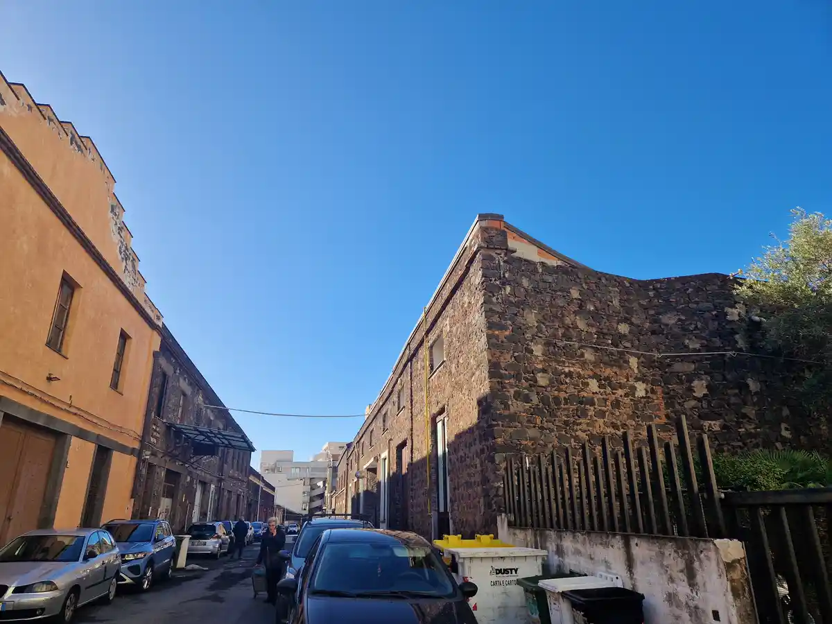 A narrow urban street with parked cars, two-story buildings with weathered facades, and a clear blue sky. A person is…