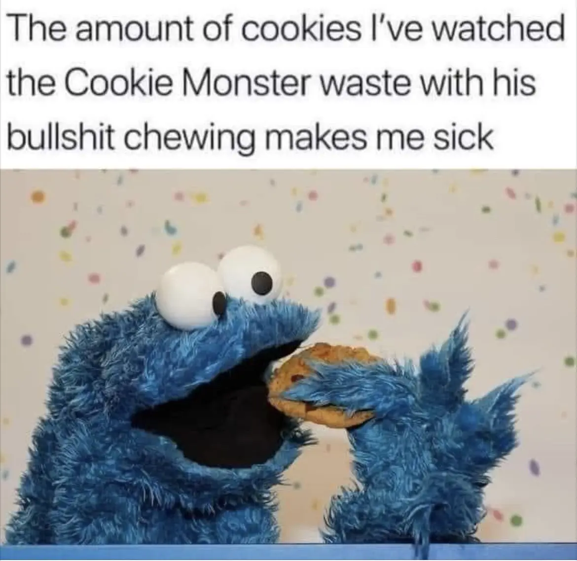 A blue, furry Cookie Monster puppet is shown mid-chew, holding a chocolate chip cookie in his hand as he eats it. The…