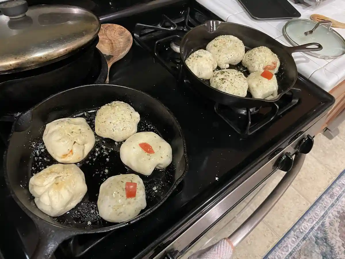 Two cast-iron skillets sit on a stovetop, each containing five round, doughy food items being fried in oil. Some have small…