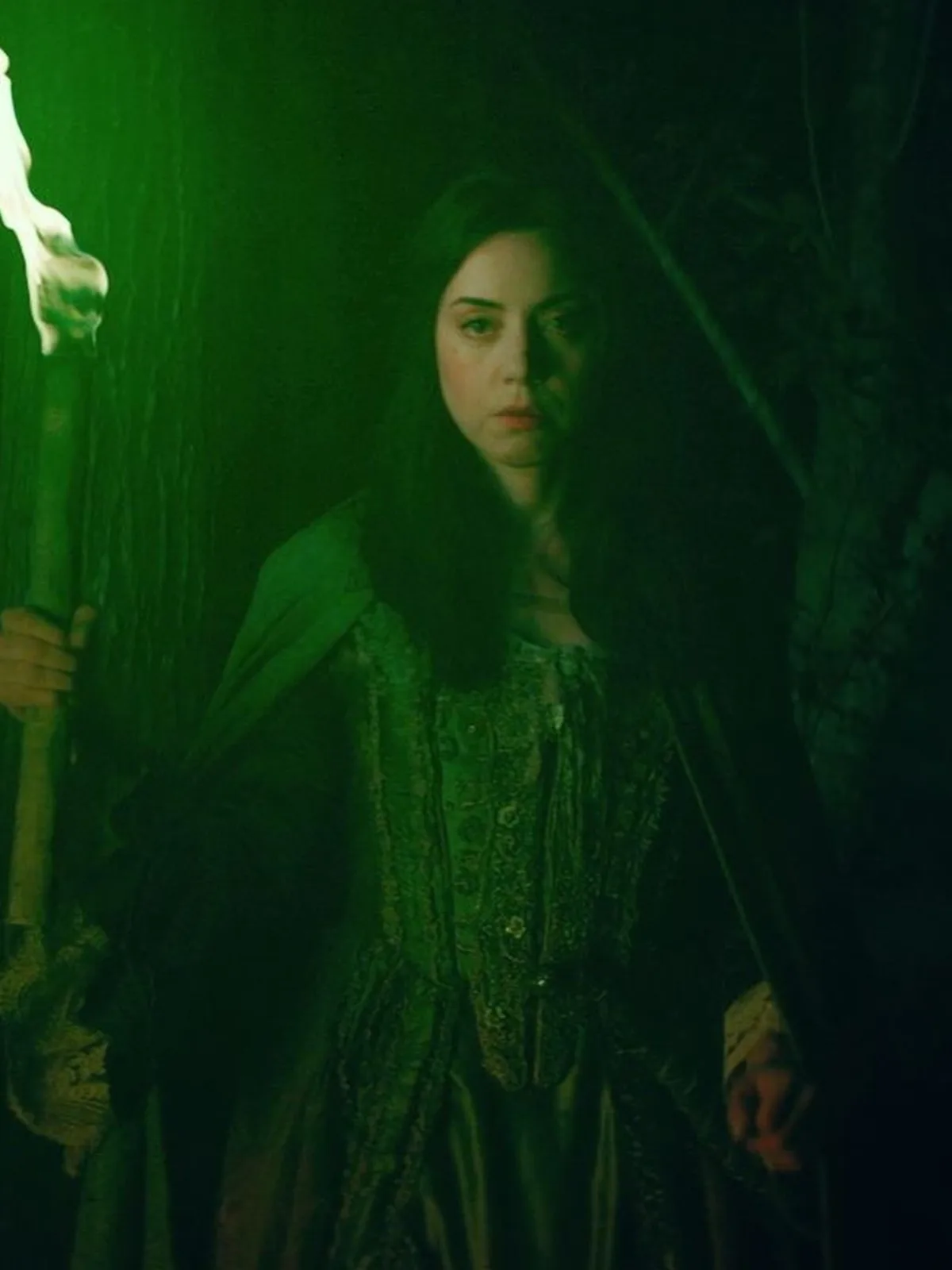 A woman with long dark hair, wearing a dark, ornate historical dress and cloak, stands in a dimly lit, green-hued forest…