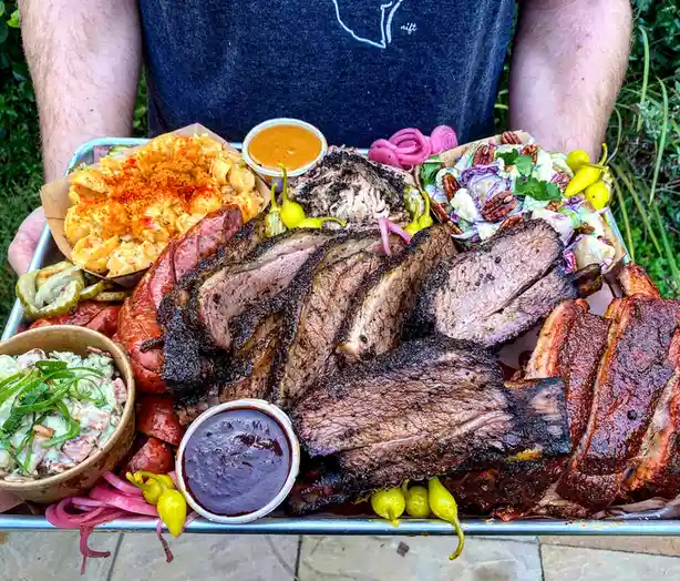 a person holds a large metal tray overflowing with a lavish barbecue spread, featuring sliced brisket, ribs, pulled pork…