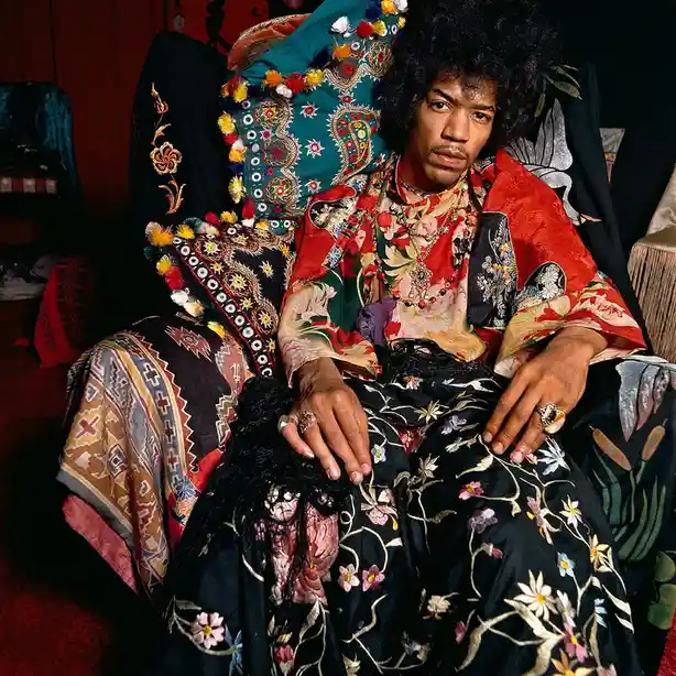 A man with an afro and mustache sits in a chair surrounded by colorful, patterned textiles and pillows. He wears a red…