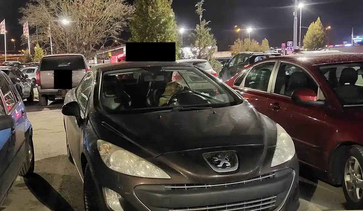 A nighttime photo of a dark gray Peugeot car parked in a crowded lot, with a person in a camouflage-patterned jacket sitting…