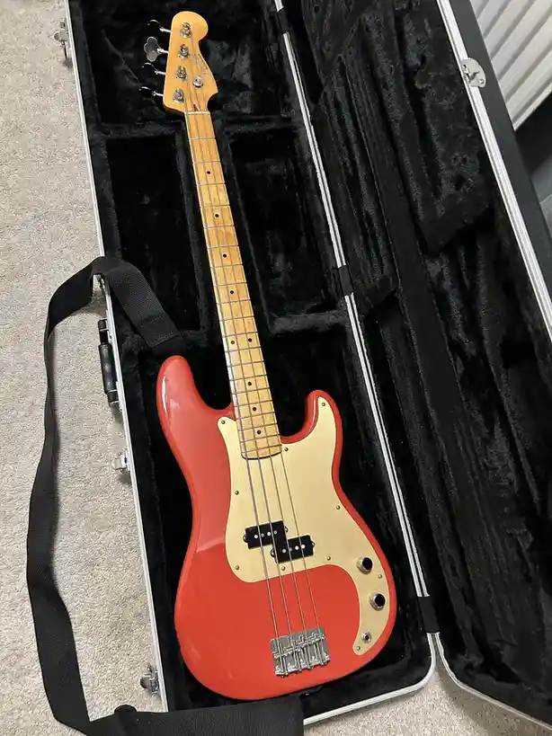 A red and natural wood-finish Fender-style bass guitar rests inside an open, black-lined hard case on a beige carpet. The…