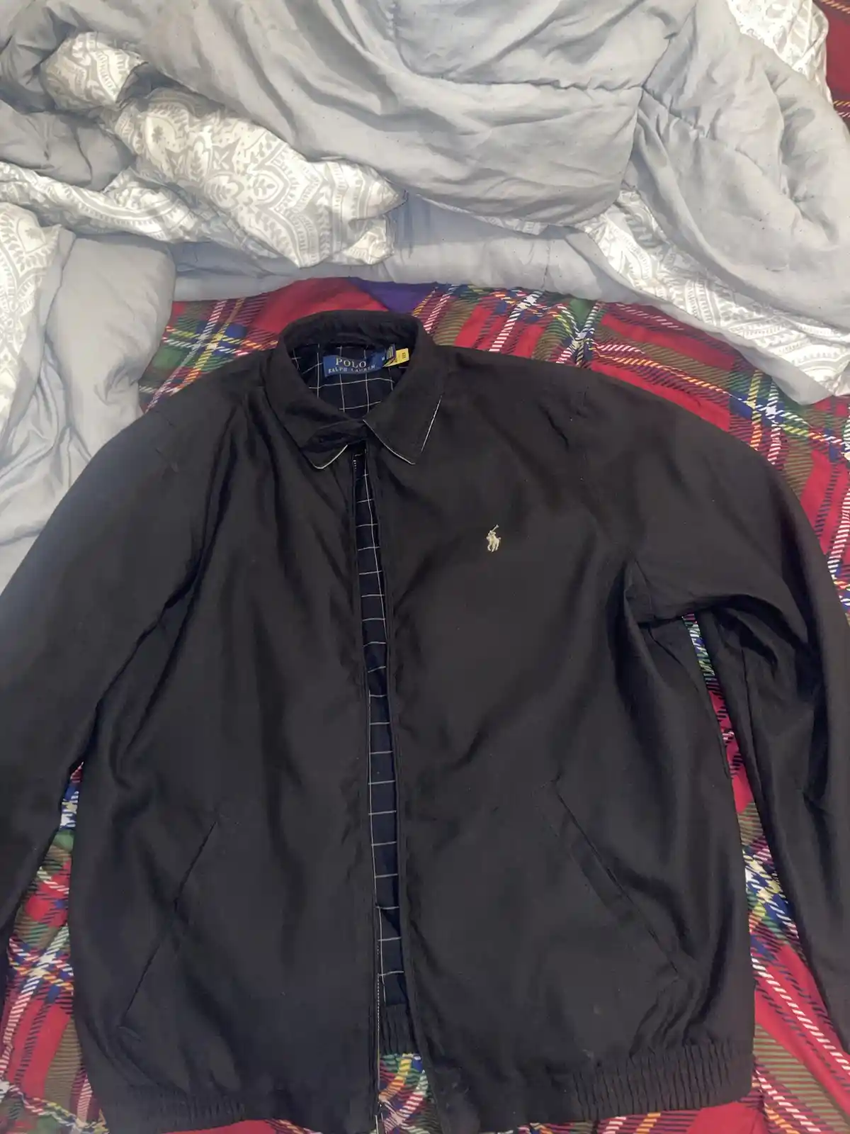 A black Ralph Lauren jacket with a small polo player logo on the left chest is laid open on a colorful plaid bedspread…