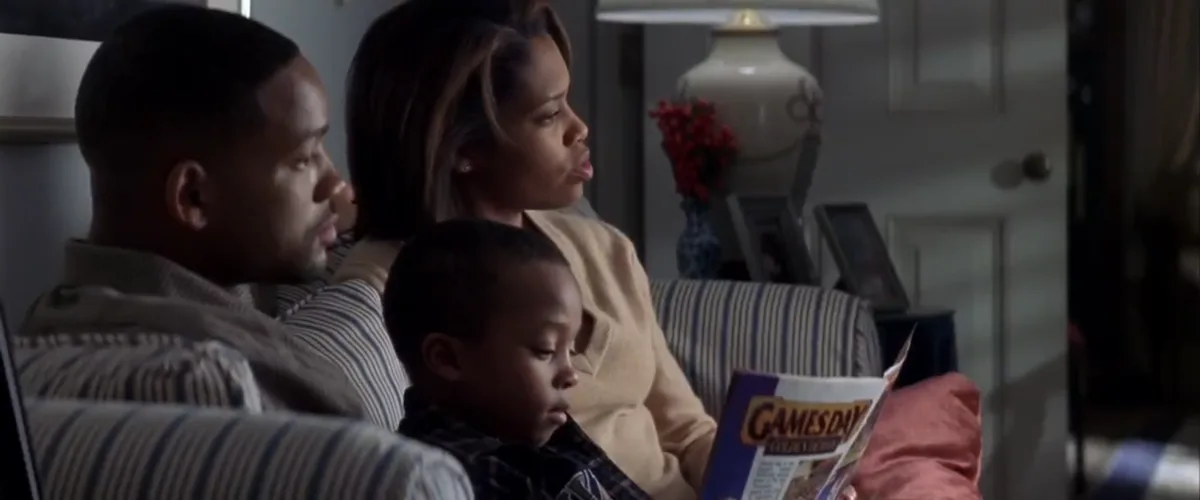 A still from a movie shows a family of three sitting on a striped couch in a living room. The father and mother are looking…