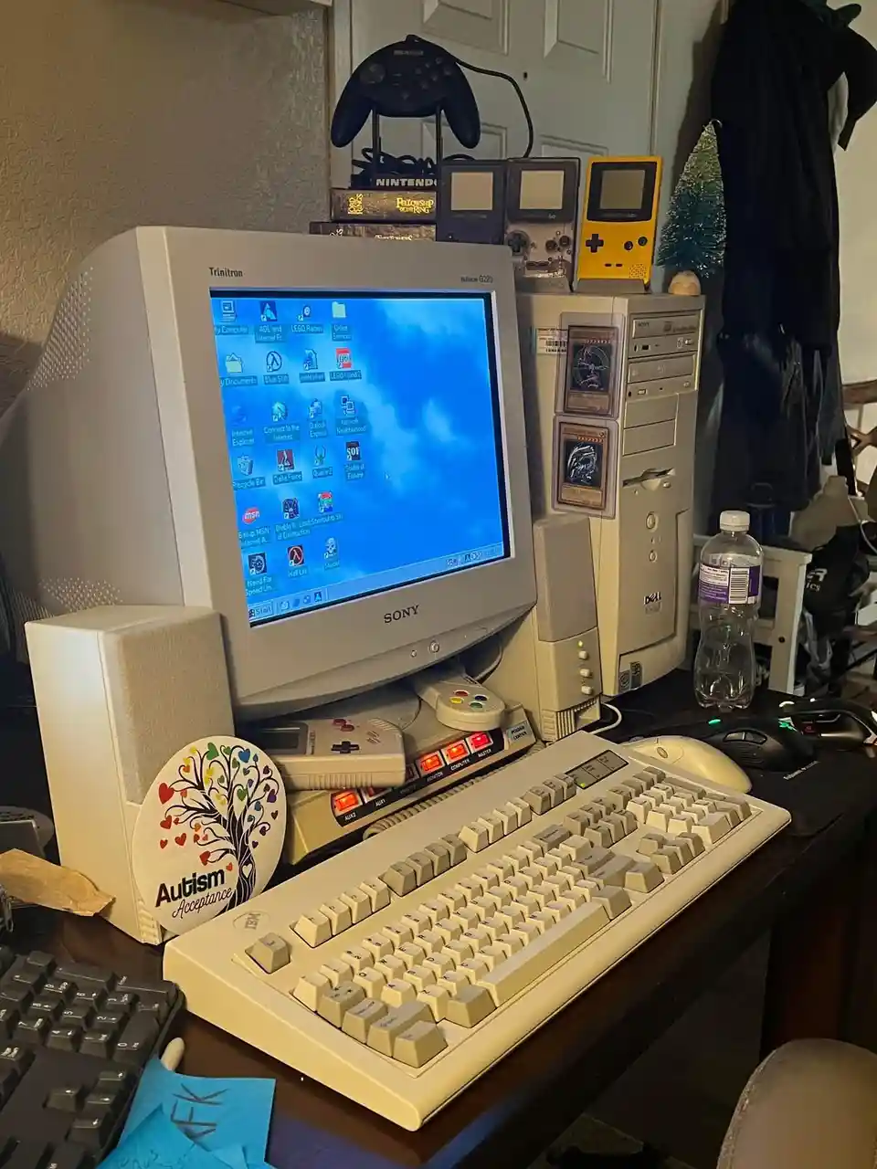A cluttered retro computer desk features a beige Sony Trinitron monitor displaying a Windows XP desktop with game icons, a…