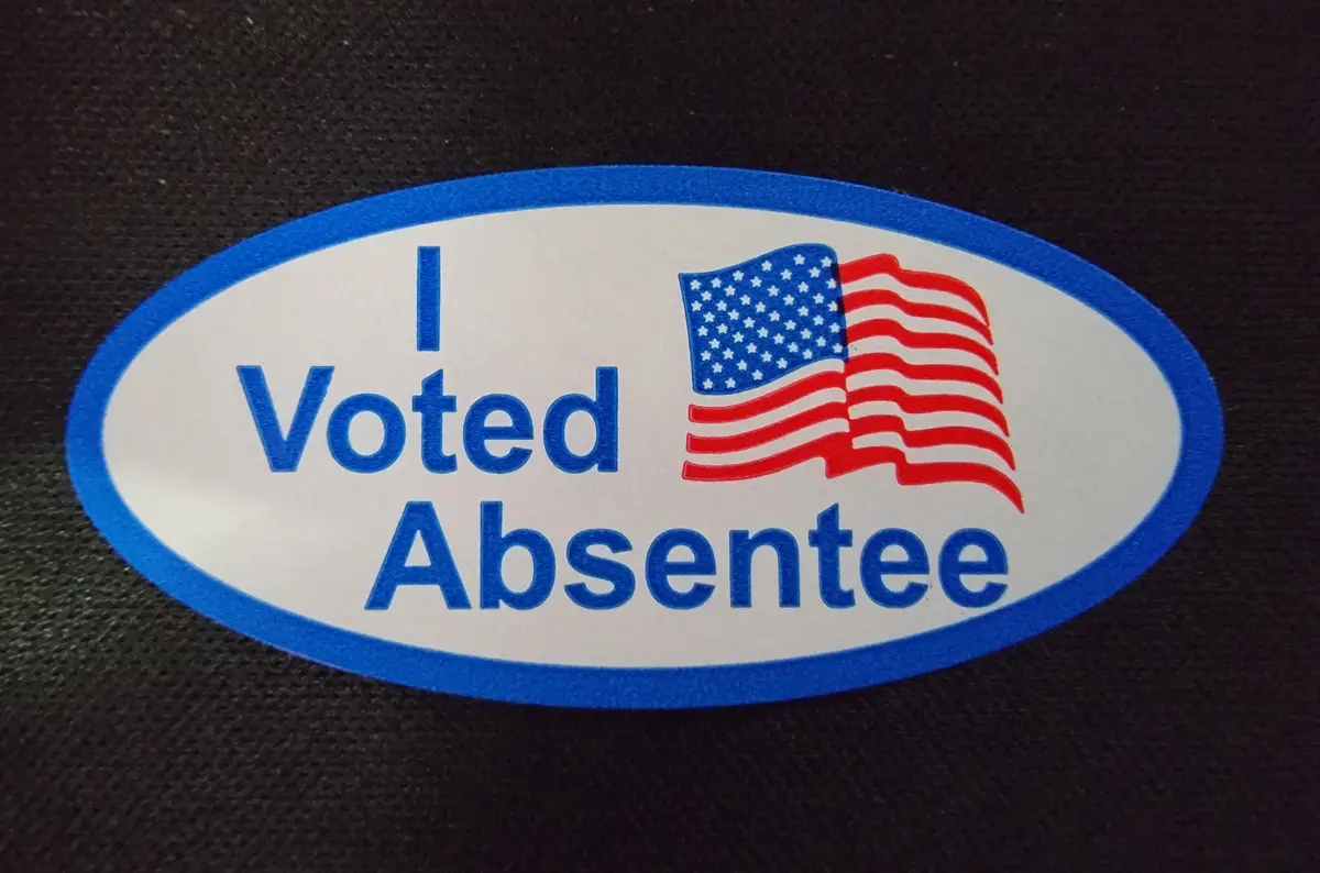 A close-up of an oval sticker with a blue border, white background, and blue text reading 'I Voted Absentee' next to a small…