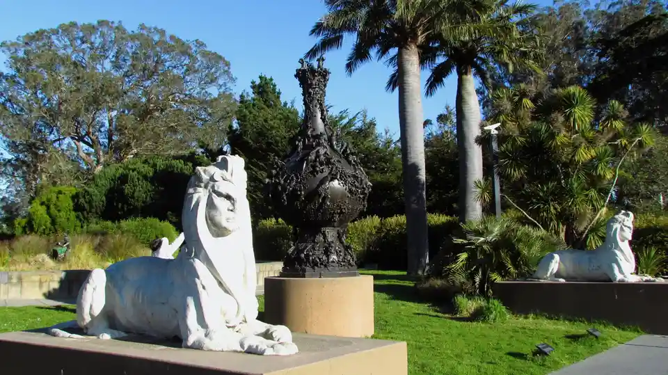 Two white sphinx statues flank a central black ornate sculpture in a sunny garden with palm trees and greenery. A person in…