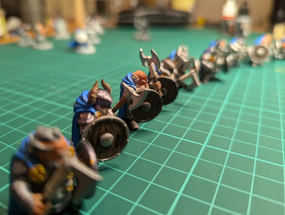 A row of miniature fantasy warrior figurines, painted in blue and silver armor with shields and axes, are lined up on a…