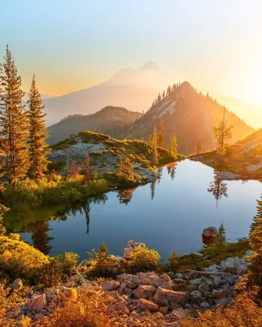 A serene mountain landscape at sunrise or sunset, featuring a calm alpine lake reflecting the sky and surrounding peaks…