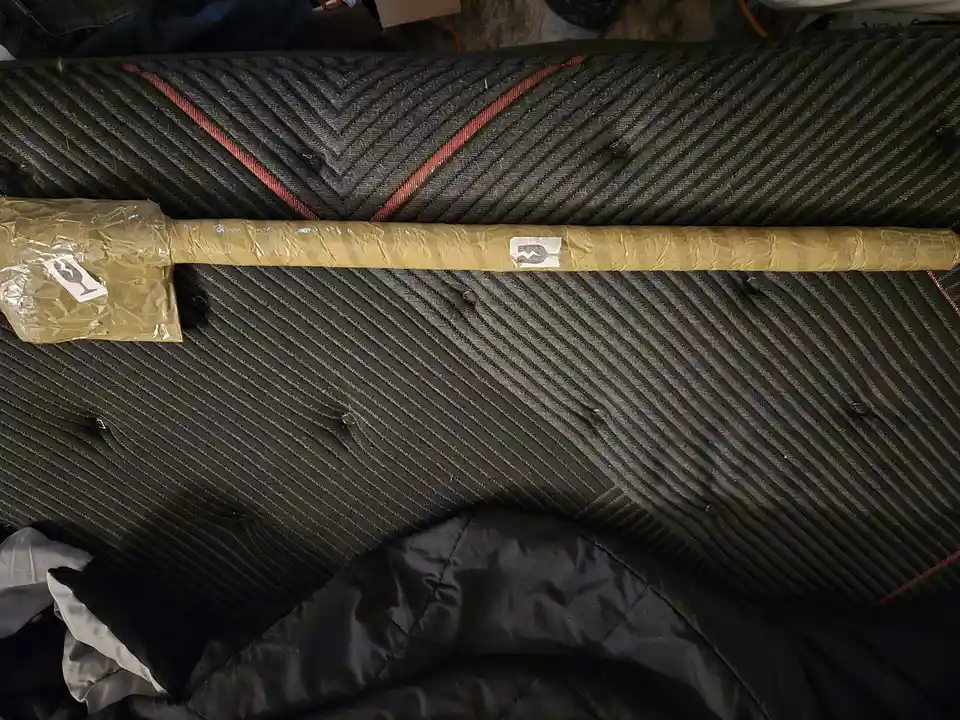 A long, cylindrical object wrapped in brown paper and clear plastic tape lies horizontally on a dark, textured fabric…