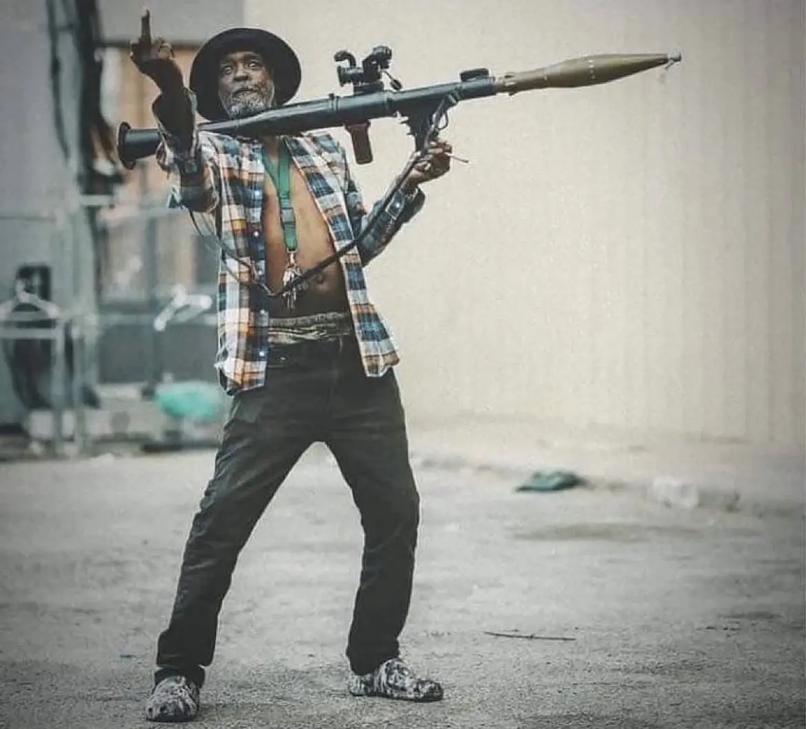 A man in a plaid shirt, hat, and patterned shoes stands in an urban alley holding a rocket launcher over his shoulder while…