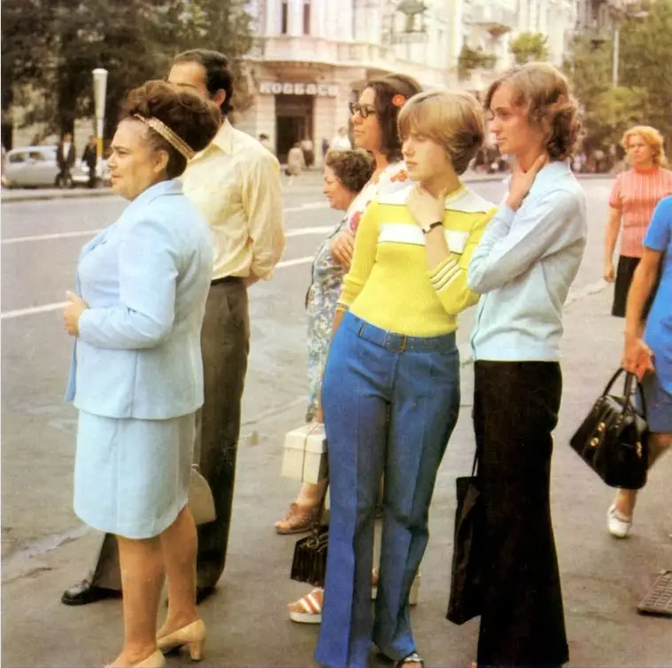 A vintage photo shows a group of people standing on a city street, with a woman in a light blue suit looking sideways with a…