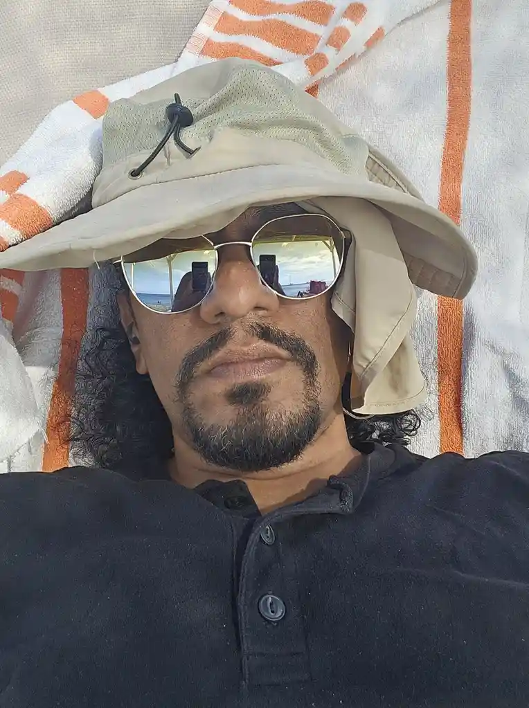 A man with a goatee and long dark hair lies on a beach chair under an orange and white striped towel, wearing a beige bucket…