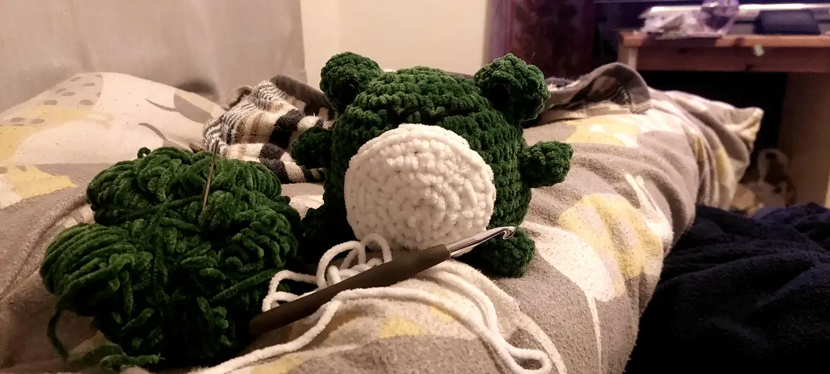 A green crocheted frog plushie with a white snout sits on a patterned blanket next to a ball of green yarn with a needle…