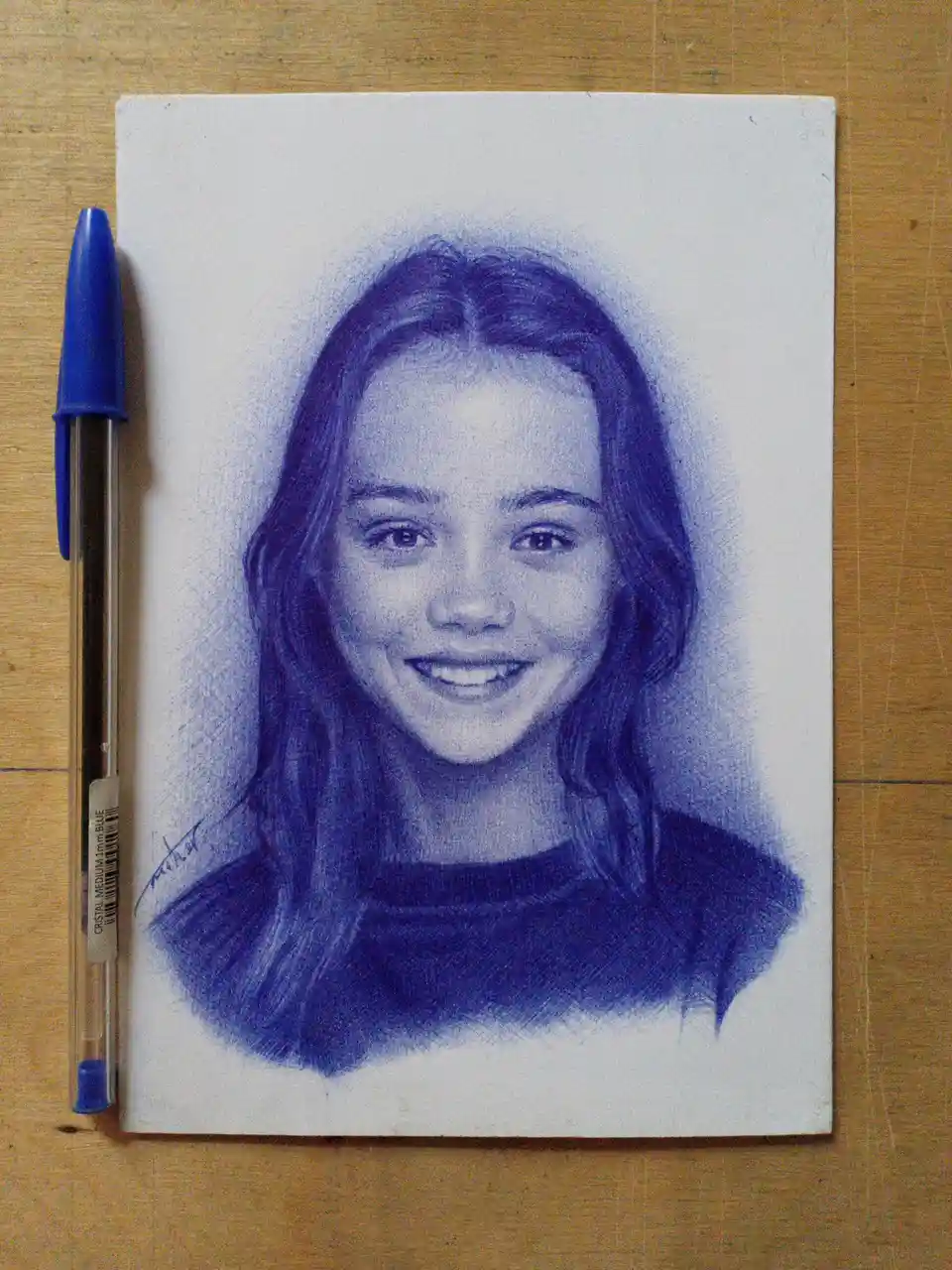 A detailed portrait drawing of a smiling young woman with long hair, rendered entirely in blue ink on white paper, is shown…