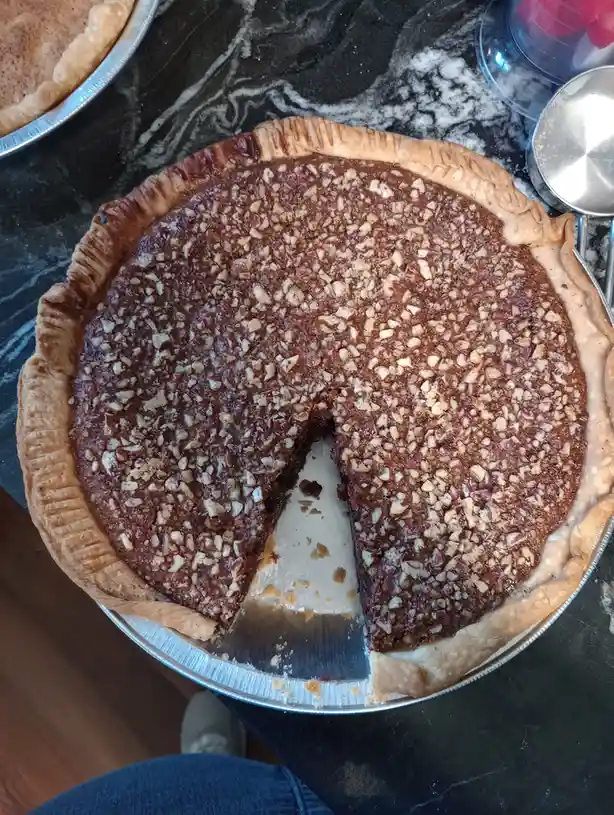 An overhead photo shows a pecan pie in a metal pie tin on a dark marble countertop, with one slice missing and crumbs…