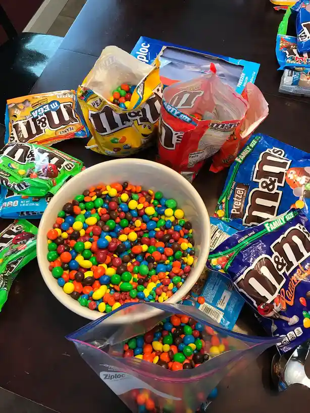 A white bowl overflowing with colorful M&M's sits on a dark table, surrounded by numerous opened and unopened bags of…