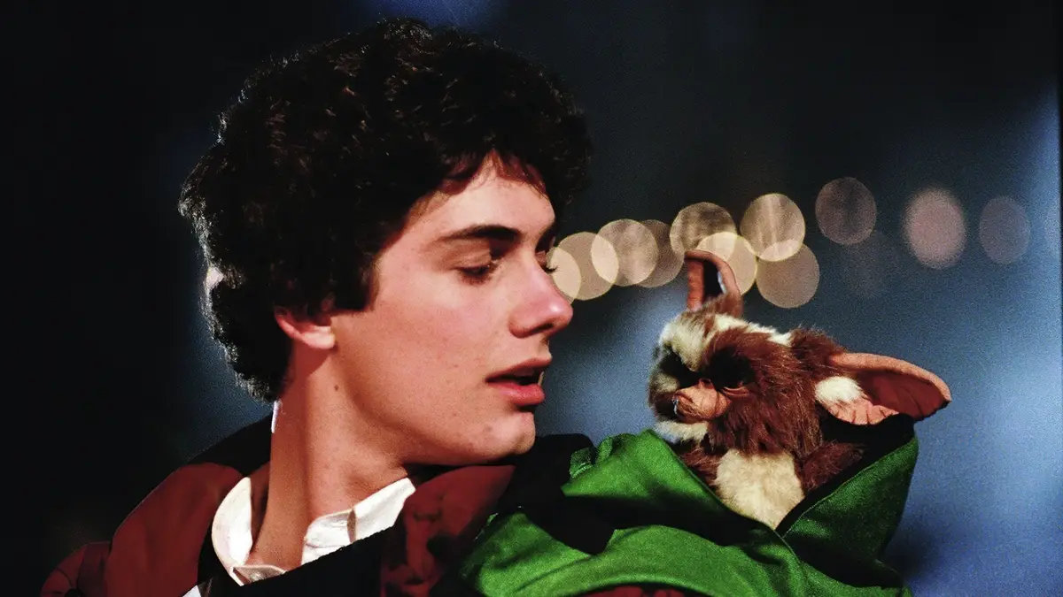 A young man with dark curly hair, wearing a red and black jacket over a white shirt, holds a small brown and white furry…