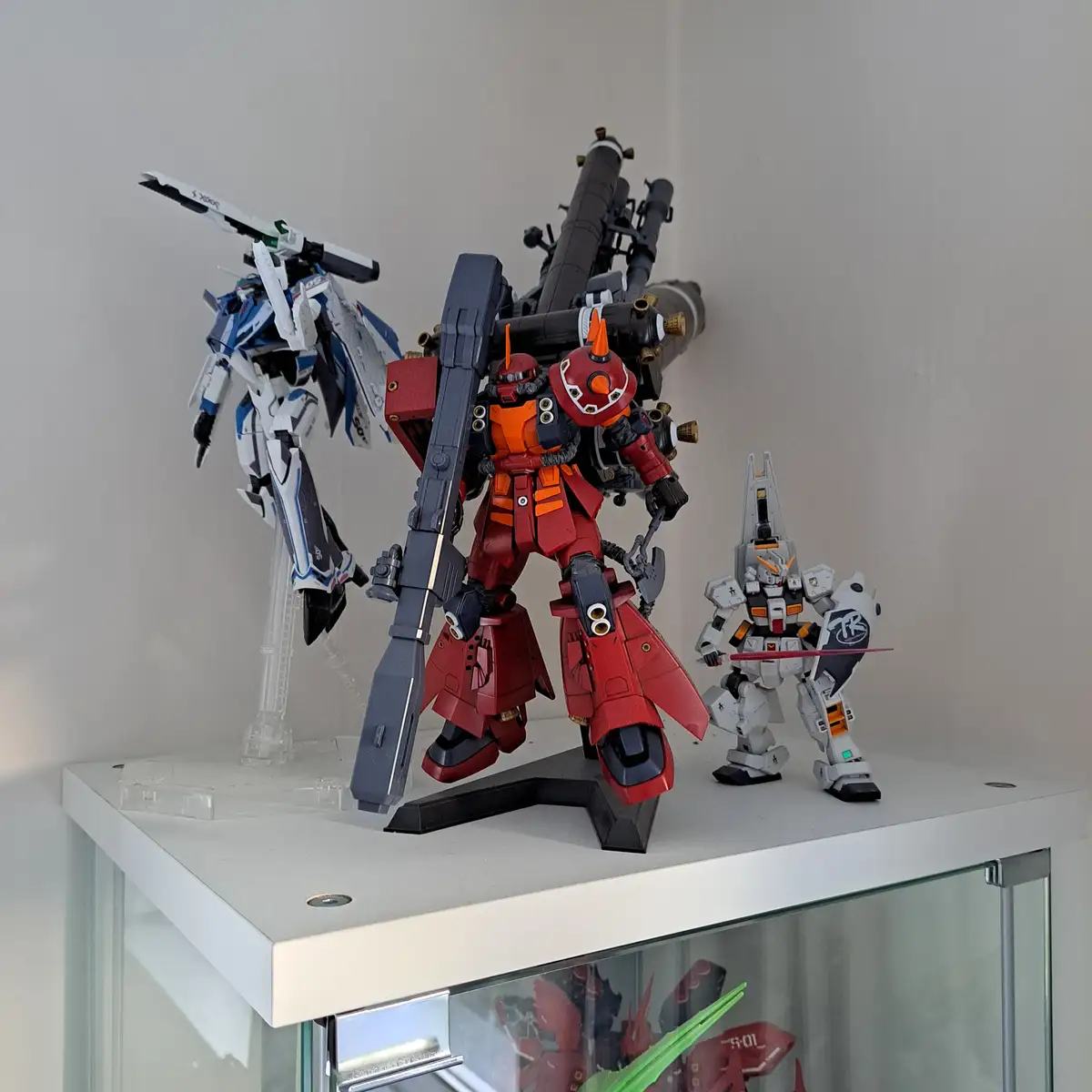 Three detailed robot model figures, resembling Gundams, are displayed on a white shelf. The central, largest figure is red…