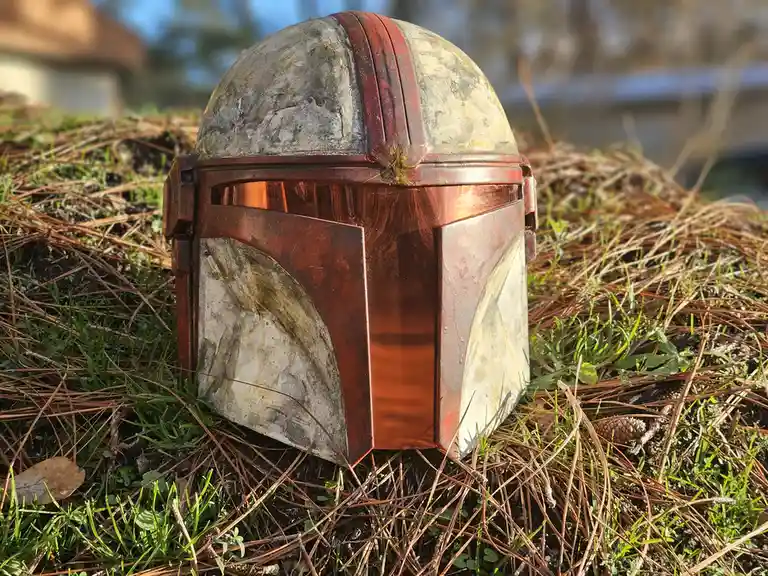 A weathered, red-and-white Mandalorian helmet from Star Wars rests on a bed of pine needles and grass outdoors. The helmet…