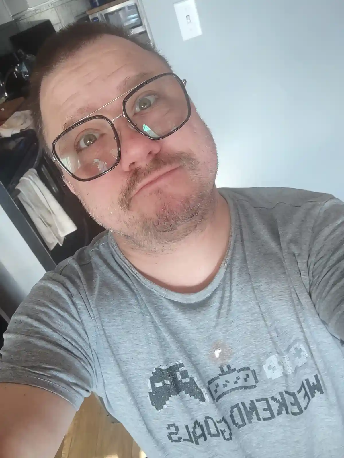 A man with large glasses and a mustache takes a selfie in a kitchen, looking upward with a smug expression. His gray t-shirt…