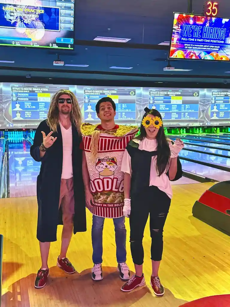 Three people in costumes pose for a photo at a bowling alley. The person on the left wears a long wig and sunglasses, making…