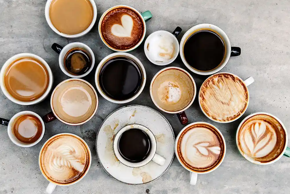 An overhead shot displays a scattered collection of coffee cups on a gray surface, featuring various coffee types from black…