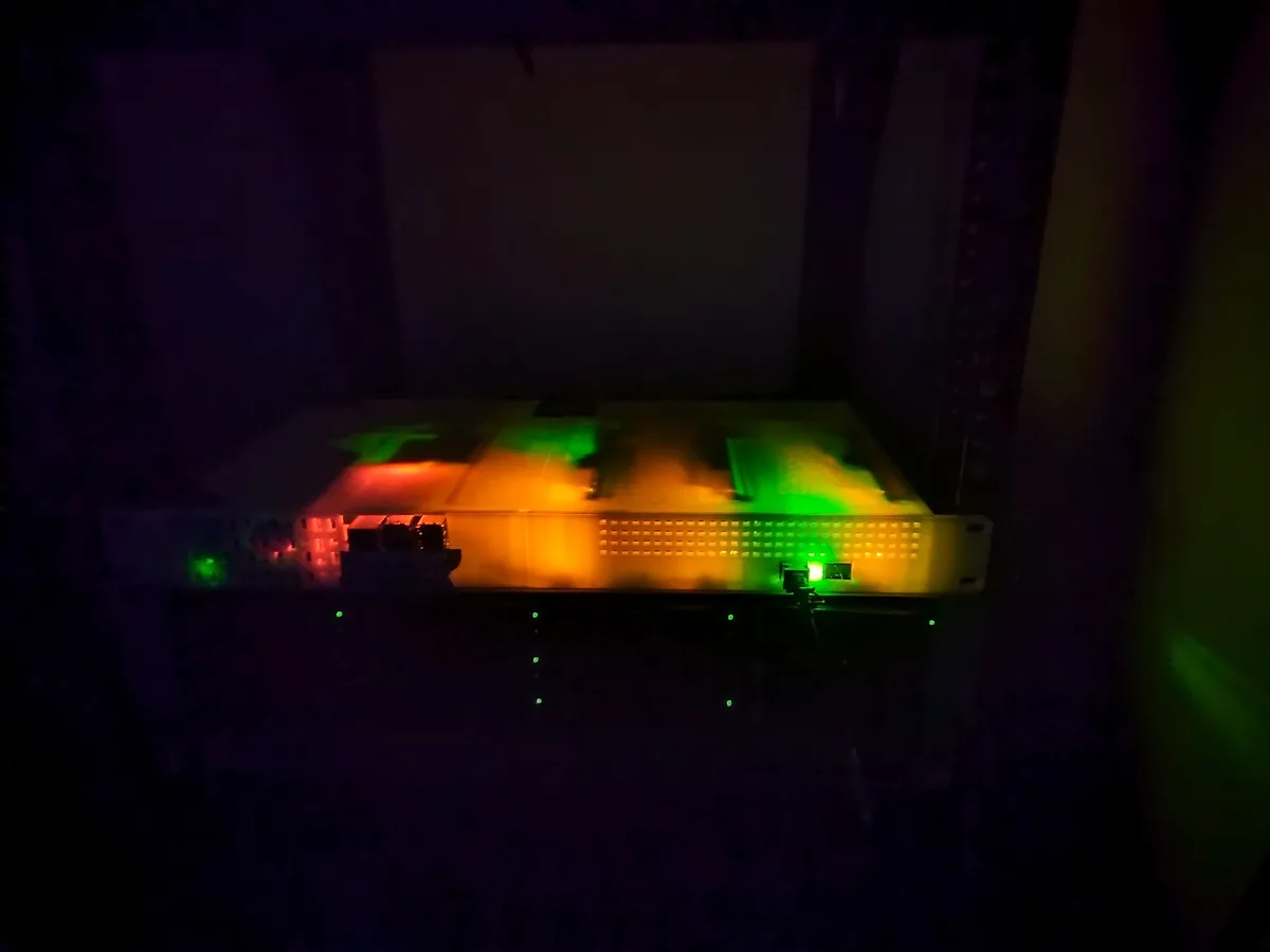 A server rack unit in a dark room is illuminated by colorful, blurry lights resembling a disco, with green and orange hues…