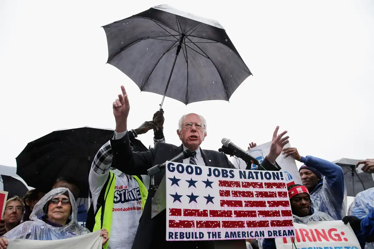 A man in a suit and glasses speaks at a rally under a black umbrella held by a supporter, surrounded by people in rain gear…