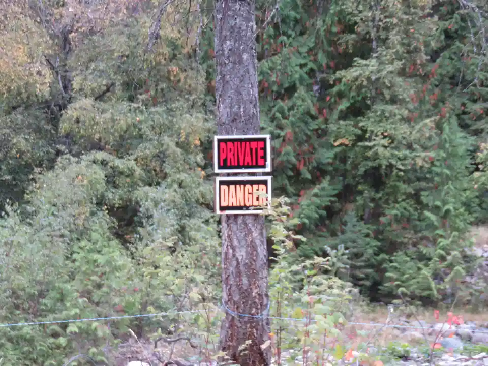 Two rectangular signs with bold red text reading 'PRIVATE' and 'DANGER' are taped vertically to a tree trunk in a forested…