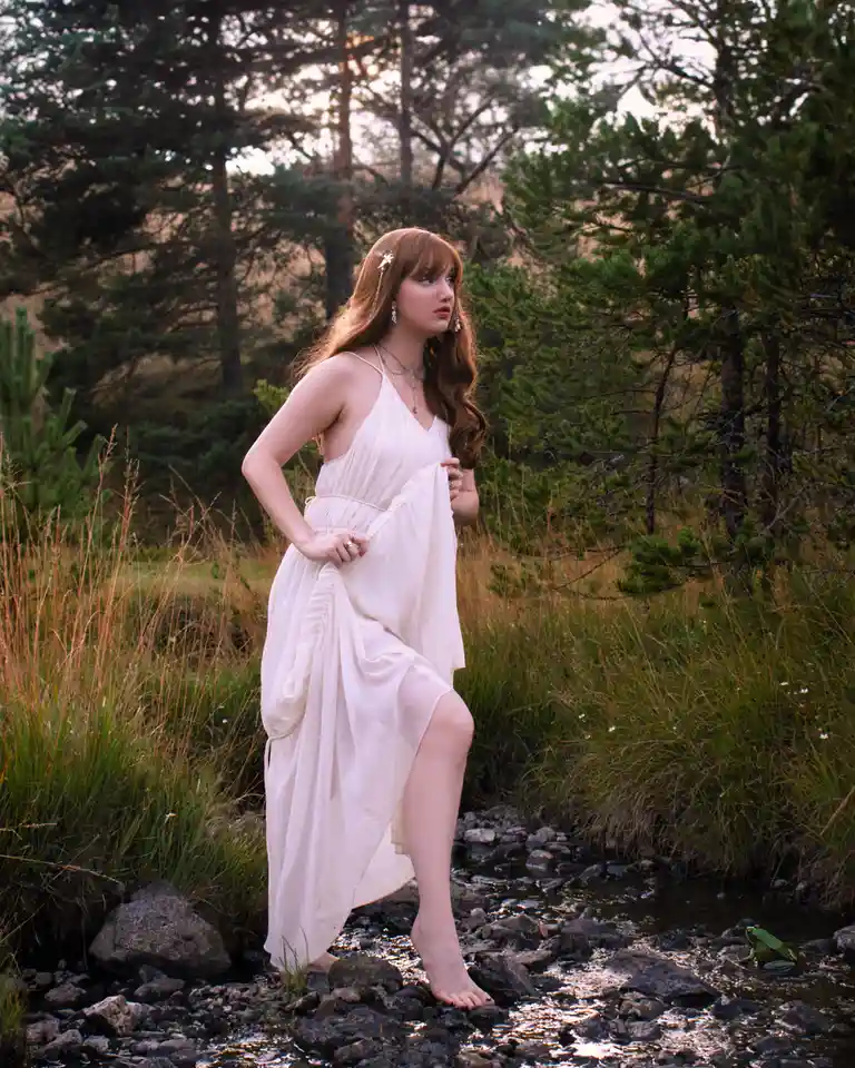 A woman in a flowing white dress stands barefoot in a shallow stream surrounded by tall grass and pine trees, looking away…