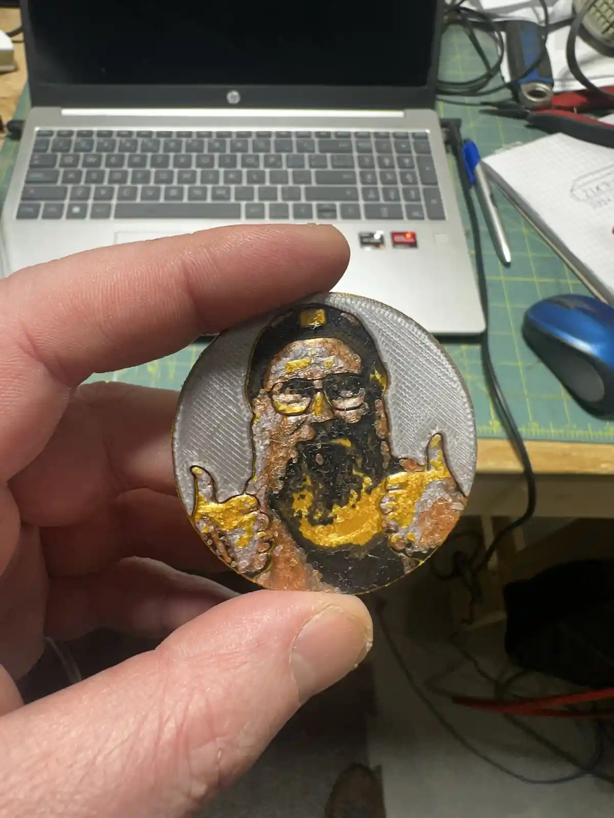 A hand holds a circular, 3D-printed coin featuring a crude, gold-and-black painted portrait of a man with glasses and a…