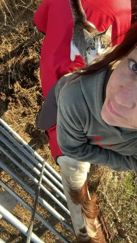 A person with red hair and freckles, wearing a grey hoodie and red jacket, takes a selfie outdoors near a metal fence. A…