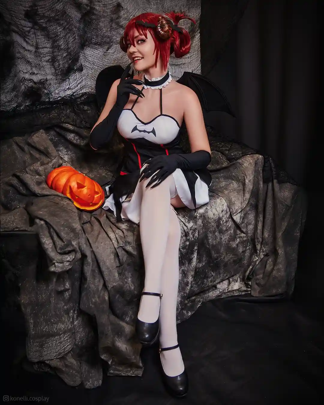 A woman with red hair styled in pigtails and wearing a black and white bat-themed costume with bat wings, black gloves, and…