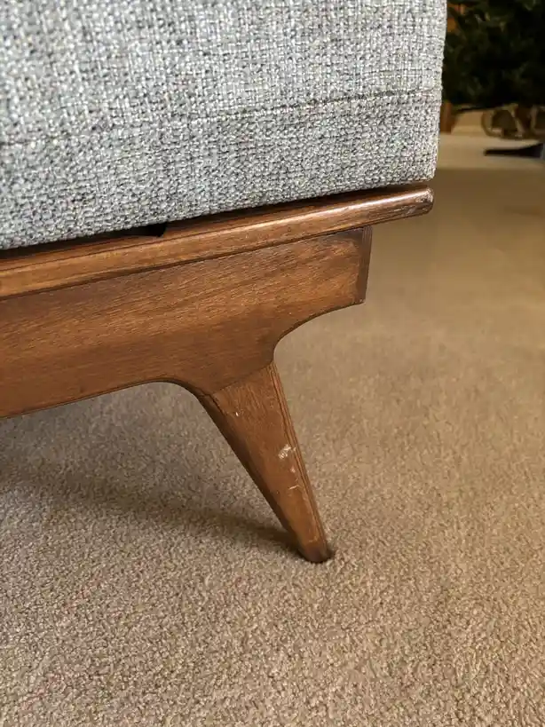 A close-up photo shows the wooden leg of a mid-century modern style couch with light blue textured fabric, standing on beige…
