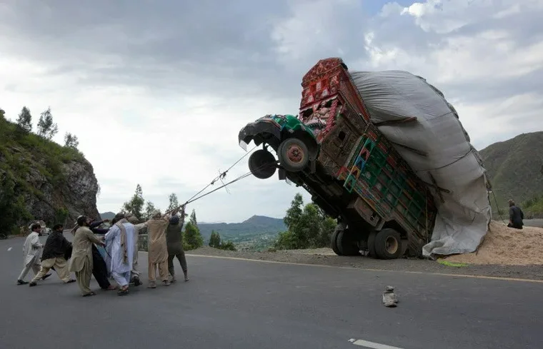 A group of men are pulling a large, heavily loaded truck that is tilted at a steep angle on a mountain road, using a rope…